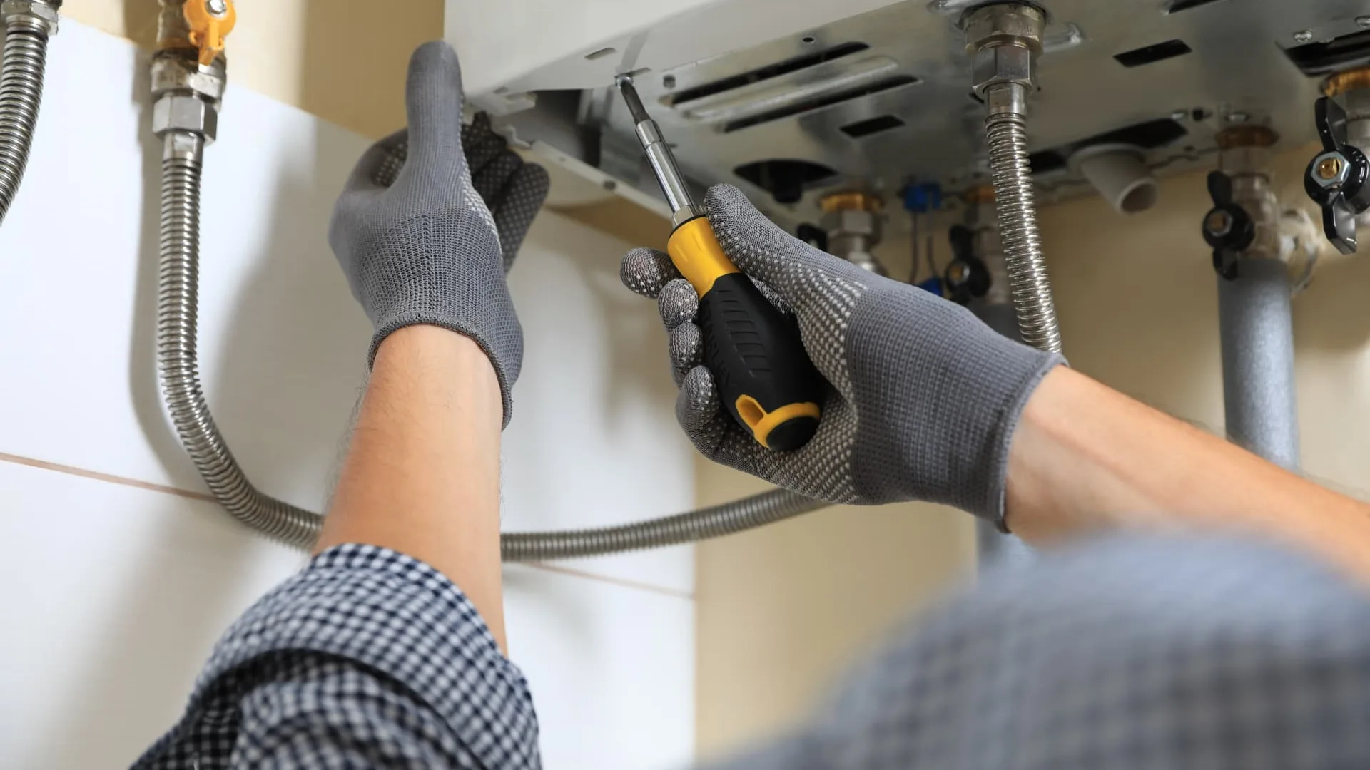 Gloved engineer using a screwdriver on the underside of a wall-mounted boiler, illustrative boiler servicing photo