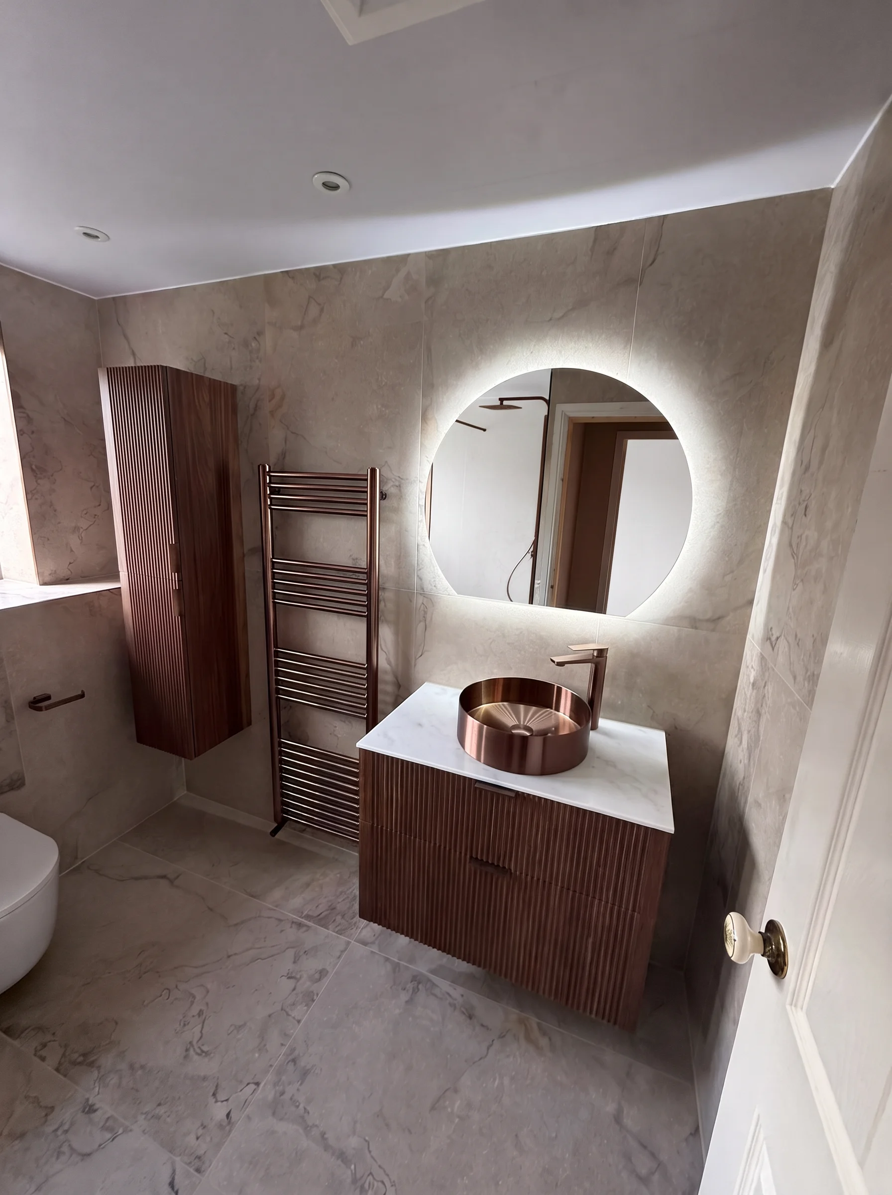 DPB bathroom with copper vessel basin on a fluted walnut vanity, backlit round mirror and copper towel radiator